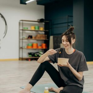A smiling woman sitting on a mat enjoying a healthy meal indoors after exercise.
