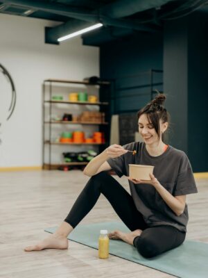 A smiling woman sitting on a mat enjoying a healthy meal indoors after exercise.