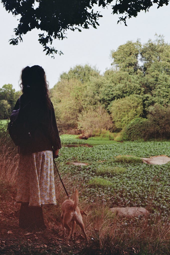 A woman walks her dog by a lush riverside in autumn, embracing nature's tranquility.